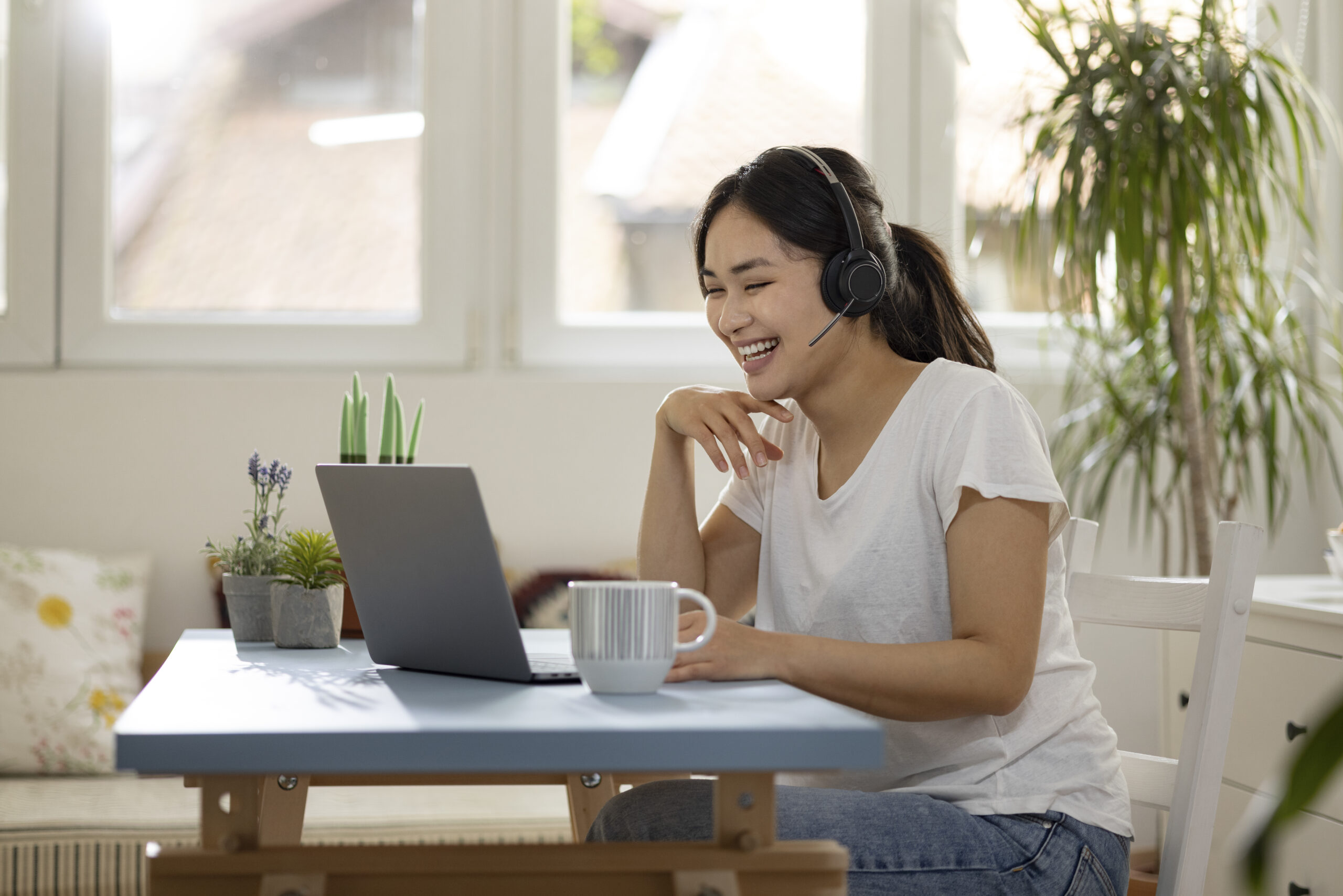 Customer support contact center female employee of Mongolian ethnicity working remotely from home, using a laptop, wearing a headset and resolving a customer’s issue with a smile