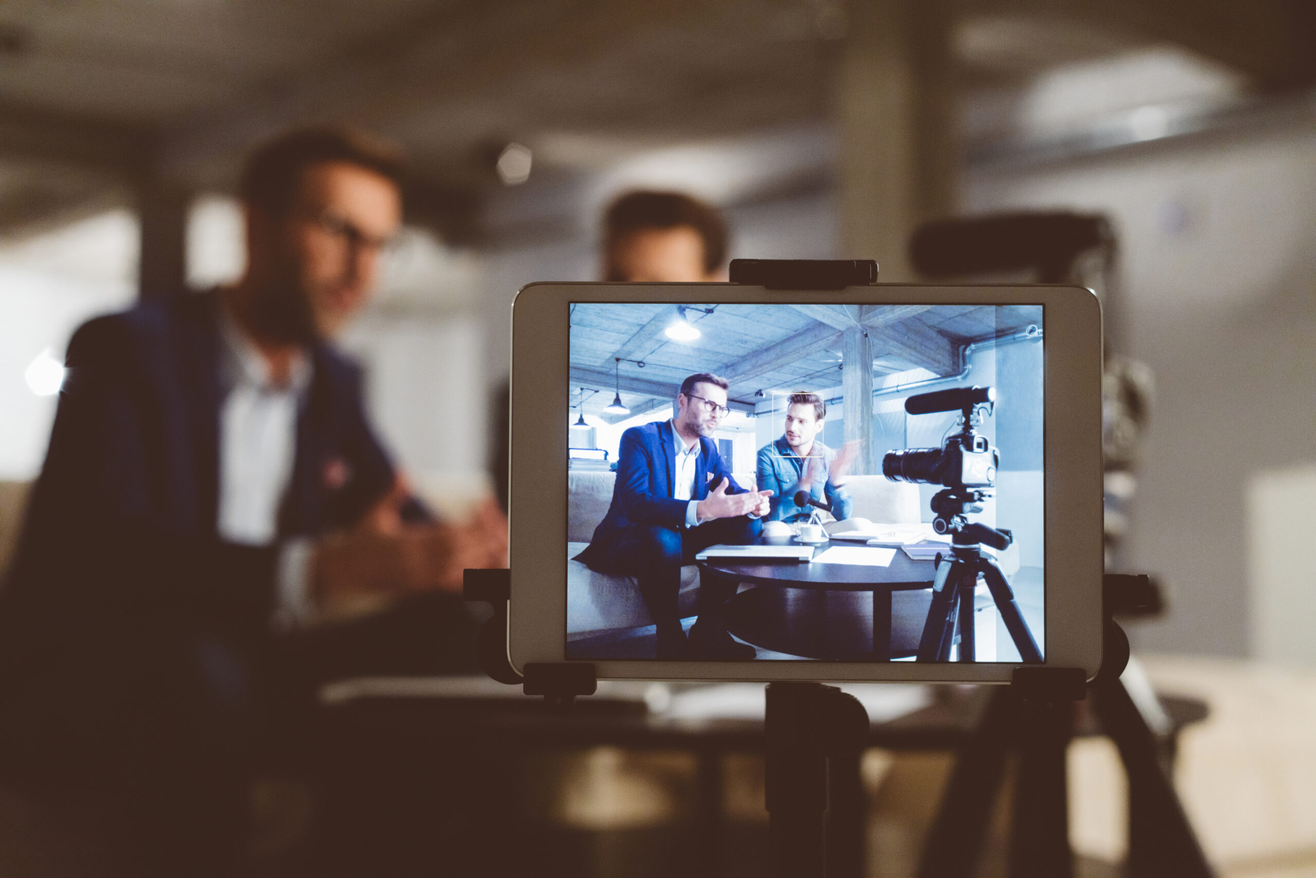 Two bloggers on digital tablet screen. Businessman with male guest recording a video blog on camera. Behind the scenes of a business vlog.