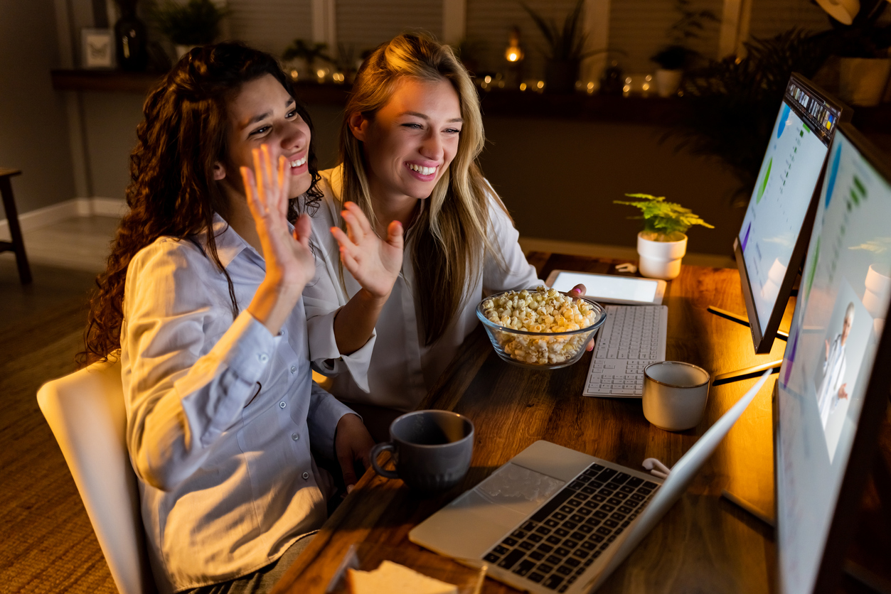 Shot of two joyful business people sitting together at desk, waving at webcam at the beginning of a video conference call at night.