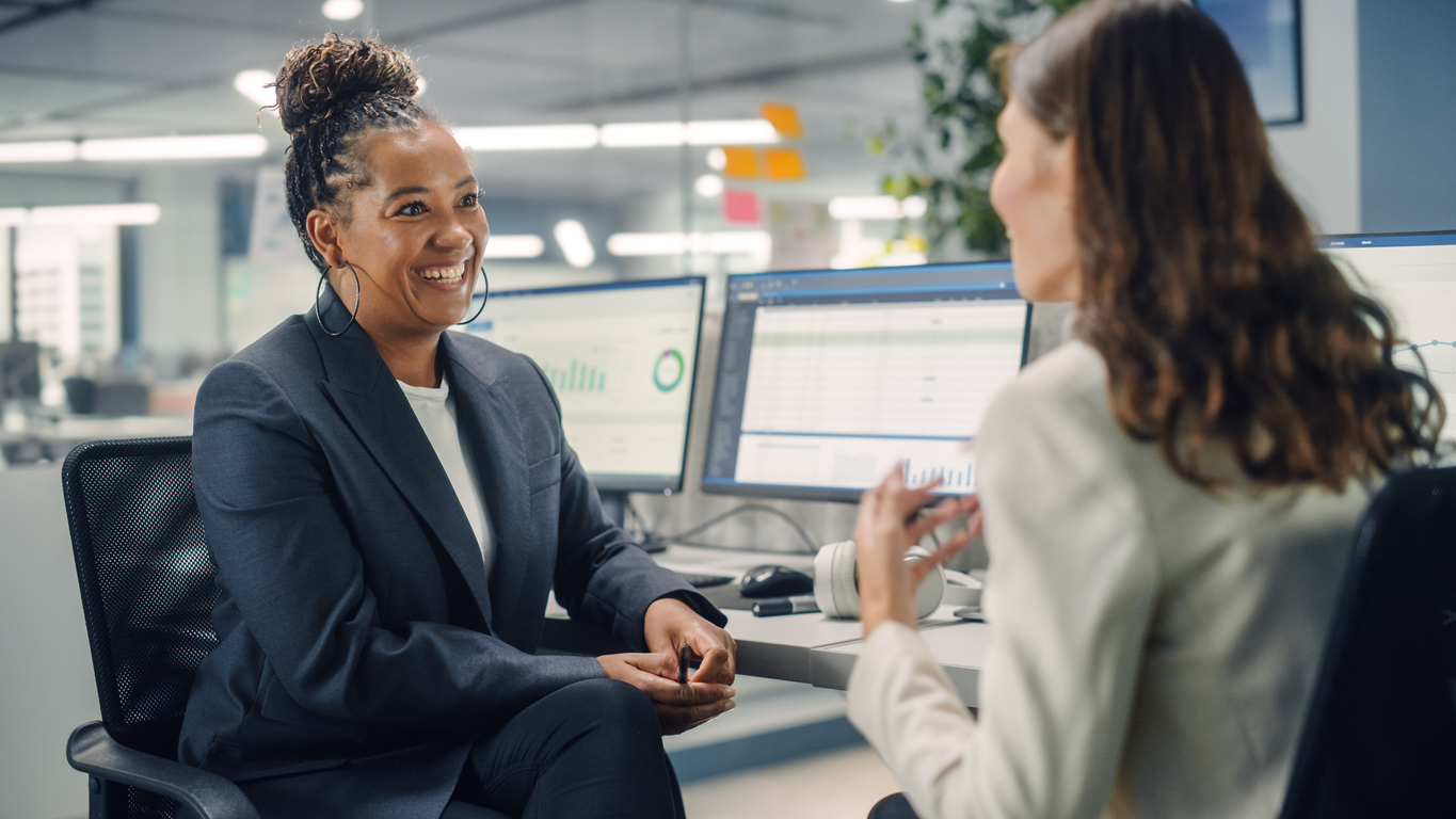 Two Female Colleagues Fondly Talk to Each Other, Laugh and Smile while Working on Computers in Diverse Modern Business Office. Experienced Manager and Young Employee Discuss a Fun Analytical Project.